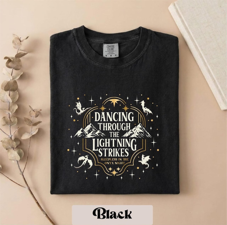 A black t-shirt with a graphic design featuring a mountain range and stars, along with the text "DANCING THROUGH THE LIGHTNING STRIKES" and "SLEEPY NIGHT".