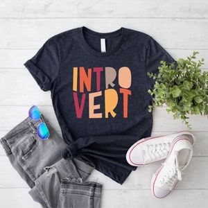 May include: A dark blue t-shirt with the word "INTROVERT" printed in colourful letters. The letters are in a retro style font and are arranged in a vertical stack. The shirt is laid flat on a white wooden surface with a pair of blue sunglasses and a pair of white trainers.