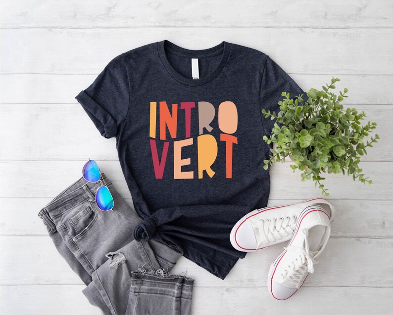May include: A dark blue t-shirt with the word "INTROVERT" printed in colourful letters. The letters are in a retro style font and are arranged in a vertical stack. The shirt is laid flat on a white wooden surface with a pair of blue sunglasses and a pair of white trainers.
