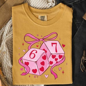 a yellow t-shirt with a graphic design of two pink dice with the number 6 on them, surrounded by hearts and ribbons.