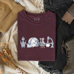 May include: Berry-colored crew neck t-shirt featuring a whimsical illustration of geese in a medical setting. The design includes a goose doctor, a CT scanner, and other medical equipment. The shirt is surrounded by a tan beanie, jeans, and boots.