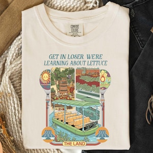 a white t-shirt with a colorful graphic design depicting a boat and various elements, along with the text "GET IN LOSER WE'RE LEARNING ABOUT Lettuce" printed on it.