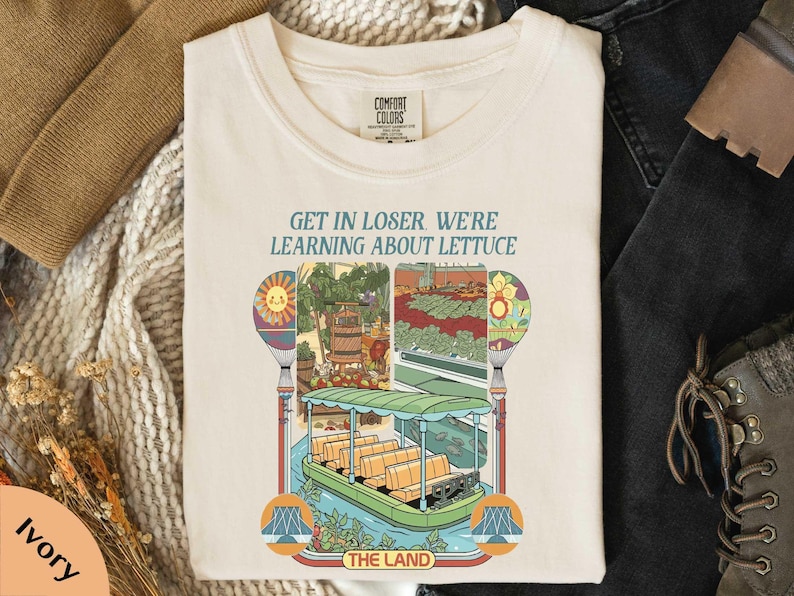 a white t-shirt with a colorful graphic design depicting a boat and various elements, along with the text "GET IN LOSER WE'RE LEARNING ABOUT Lettuce" printed on it.
