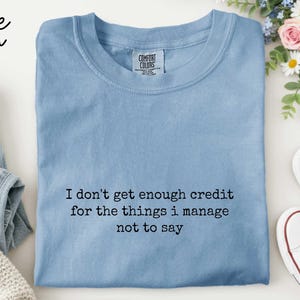 a blue t-shirt with a humorous text printed on it, along with a pair of jeans and sneakers, all arranged on a white background.