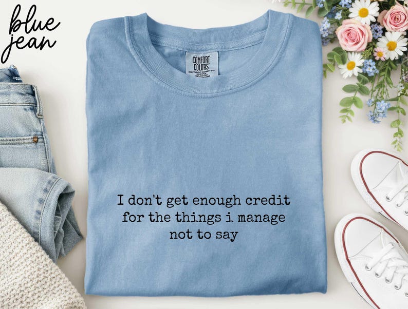 a blue t-shirt with a humorous text printed on it, along with a pair of jeans and sneakers, all arranged on a white background.