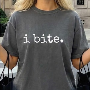 May include: Dark gray t-shirt with the text "i bite." in white. The shirt is worn by a person holding black shopping bags. The person is smiling and wearing a white skirt.