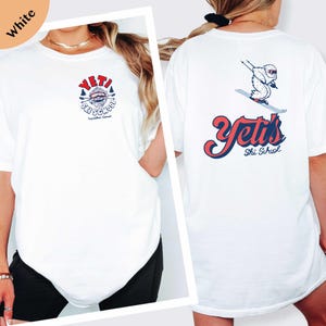 A white t-shirt with a graphic design on the front and back, featuring a snowboard and the text "YETI Ski School".