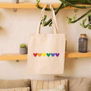 May include: A white canvas tote bag with a rainbow of hearts printed on the front. The hearts are red, orange, yellow, green, blue, indigo, and violet.