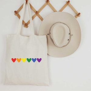 May include: A white canvas tote bag with a rainbow of hearts printed on the front. The bag is hanging on a wooden rack.