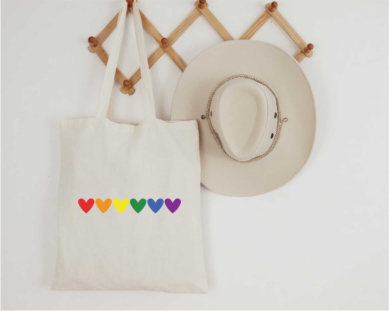 May include: A white canvas tote bag with a rainbow of hearts printed on the front. The bag is hanging on a wooden rack.