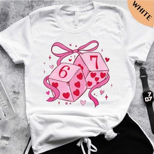 A white t-shirt with a graphic design featuring two pink dice with the number 6 and 7 on them, surrounded by hearts and stars.