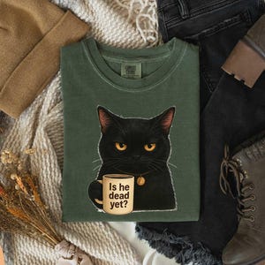 May include: Green t-shirt featuring a black cat with yellow eyes holding a mug. The mug reads "Is he dead yet?" A brown beanie, black jeans, and brown boots are also visible.