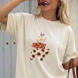 May include: An ivory short-sleeved t-shirt featuring a central graphic of three stacked animals: a cow with brown spots at the bottom, a pig with a floral pattern in the middle, and a smaller animal with a floral design at the top. The text ivory is visible.