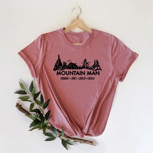 K&ouml;nnte beinhalten: Ein pinkfarbenes T-Shirt mit einer schwarzen Grafik einer Bergkette und dem Text "Mountain Man" mit den Worten "Thunder - Space - Splash - Everest" darunter.