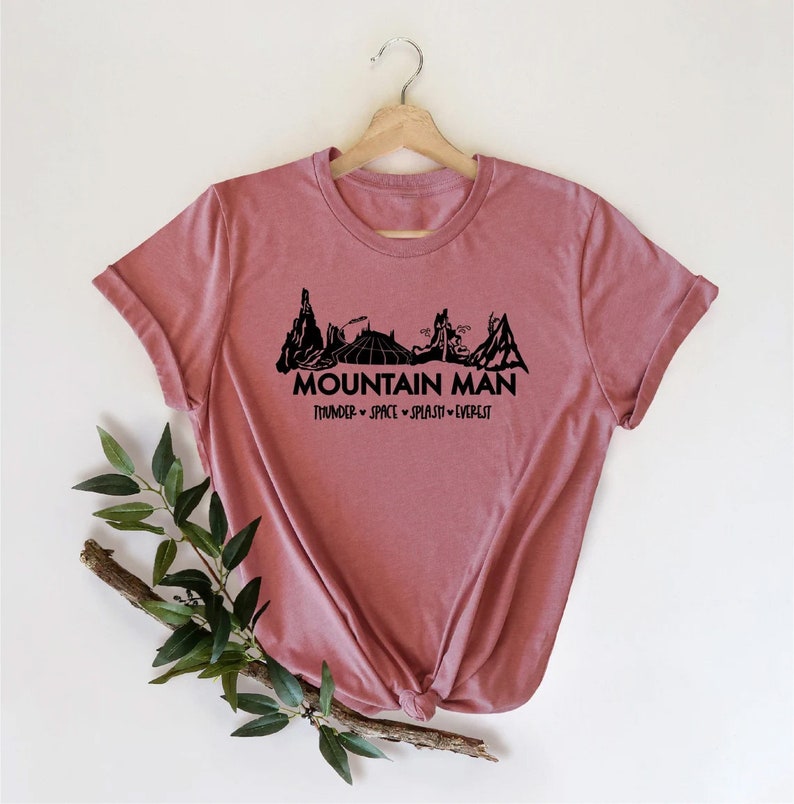 K&ouml;nnte beinhalten: Ein pinkfarbenes T-Shirt mit einer schwarzen Grafik einer Bergkette und dem Text "Mountain Man" mit den Worten "Thunder - Space - Splash - Everest" darunter.