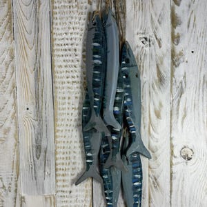 May include: Six wooden fish sculptures, painted in grey and blue, hang against a backdrop of weathered, white-painted wooden planks. The fish feature detailed fins and markings, and are tied together with twine, creating a nautical theme.