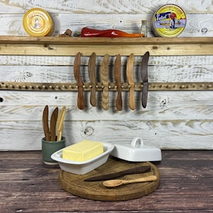 May include: A selection of wooden butter knives and a butter dish with a lid. The knives are displayed on a wooden rack and in a small green ceramic container. A wooden cutting board holds a butter knife and a small wooden spoon.