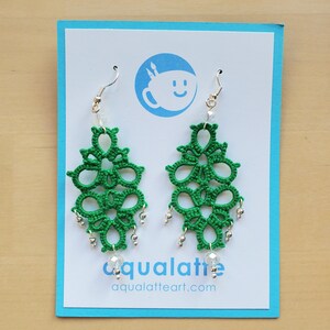 May include: A pair of green, handmade earrings with a floral design. The earrings are made of a woven material and feature silver-colored beads and clear crystal accents. The earrings hang from silver-colored hooks and are displayed on a blue card.