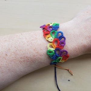 May include: A rainbow-colored bracelet crafted with intricate tatting lace techniques. The design features a repeating series of interconnected loops in vibrant shades of red, orange, yellow, green, blue, and purple, secured with thin matching thread ties.