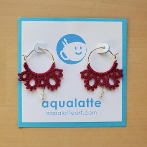 May include: A pair of burgundy hoop earrings with a delicate, lacy design. Each earring features a silver hoop and a dangling clear bead. The earrings are displayed on a blue card with the text "aqualatte" and "aqualatteart.com".