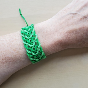 May include: A green macrame bracelet featuring an intricate, repeating scalloped pattern. The bracelet is crafted from thin, vibrant green cord and includes a matching green string tie closure that extends from one side of the woven design.