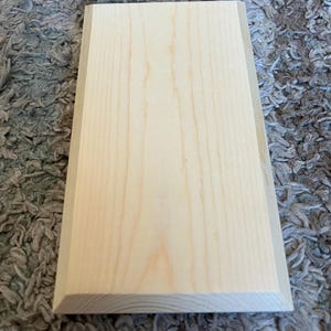 K&ouml;nnte beinhalten: Ein rechteckiges, unfertiges Holzschild mit heller, nat&uuml;rlicher Holzmaserung. Das Schild hat eine abgeschr&auml;gte Kante, die einen subtilen Rahmen um die Hauptfl&auml;che bildet. Das Holz ist glatt und etwa 20 cm lang.
