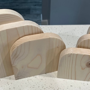 May include: A collection of unfinished, light-colored wooden shapes, possibly for crafting or decoration. The pieces have a rounded top and a flat base, with visible wood grain. The shapes vary in size, suggesting a set or collection.