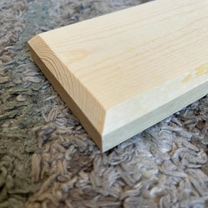 May include: A rectangular piece of unfinished light-colored wood with a beveled edge. The wood grain is visible, and the piece is resting on a textured, gray surface. The beveled edge is at a 45-degree angle.