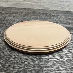 May include: A round, unfinished wooden plaque with a decorative edge. The plaque is light brown and appears to be made of solid wood. The surface is smooth and ready for painting or staining. Ideal for crafts and DIY projects.