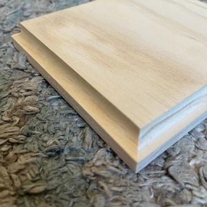 May include: Close-up of a light-colored, unfinished wooden square with a decorative edge. The wood grain is visible, and the edges are precisely cut. The item appears to be a small decorative piece or a component for a larger project.