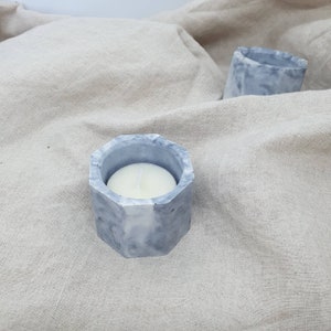 Peut inclure: Trois bougeoirs en béton gris. L'un d'eux contient une bougie blanche. Les bougeoirs sont hexagonaux et ont un aspect marbré. Le fond est un tissu de couleur neutre.