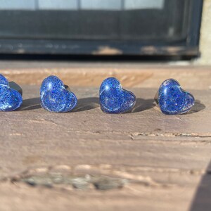 May include: Four heart-shaped stud earrings. Each earring is a translucent blue with embedded glitter. The earrings are set on a dark metal post. The earrings are displayed on a weathered wooden surface.
