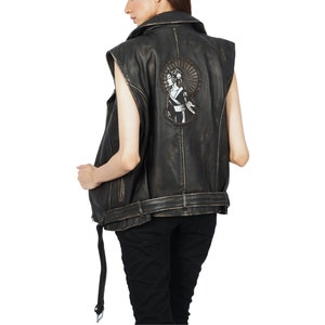 Op de afbeelding: Zwart leren vest met een verweerde look en een grote geborduurde patch op de rug met een vrouw met een waaier. Het vest heeft een riem met een gesp en een ritssluiting.