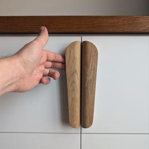 May include: A pair of wooden cabinet pulls with a rounded, slightly curved shape. The pulls are made of light brown wood and are mounted on a white cabinet door.