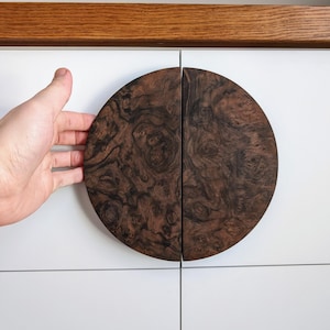 Op de afbeelding: Twee halfronde kastgrepen van donker hout met een natuurlijk houtnerfpatroon. De grepen zijn gemonteerd op een witte kastdeur.