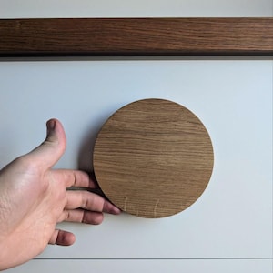 Puede incluir: Un pomo redondo de madera con un acabado marrón claro. El pomo está montado en una superficie blanca.