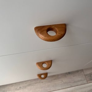 Peut inclure: Trois poignées de tiroir en bois en forme de demi-cercle avec un trou rond au centre. Les poignées sont montées sur un meuble blanc.
