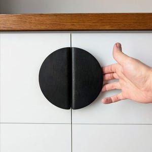 May include: Black wooden cabinet door pull with a half-circle design. The pull is mounted on a white cabinet door.