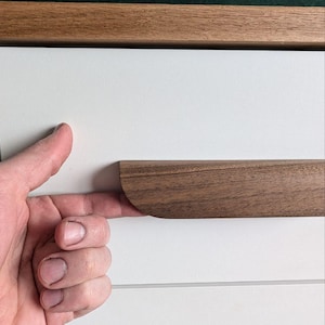 Puede incluir: Primer plano de un armario blanco con un tirador de cajón de madera oscura. El tirador del cajón tiene forma rectangular curvada. El armario tiene una moldura de madera oscura en la parte superior. El fondo es de color verde oscuro.
