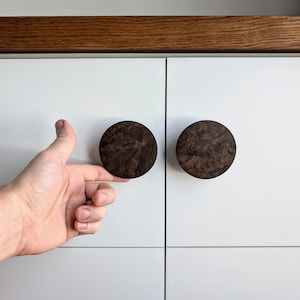 Peut inclure: Deux boutons ronds en bois foncé pour armoire, avec un motif de grain de bois. Les boutons sont montés sur une porte d'armoire blanche.
