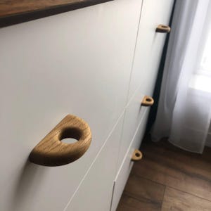 K&ouml;nnte beinhalten: Ein wei&szlig;er Schrank mit mehreren halbkreisf&ouml;rmigen Holzgriffst&uuml;cken f&uuml;r Schubladen. Jeder Griff weist eine nat&uuml;rliche Holzmaserung und eine mittige kreisf&ouml;rmige Aussparung auf und ist horizontal an der glatten, wei&szlig;en Oberfl&auml;che montiert.