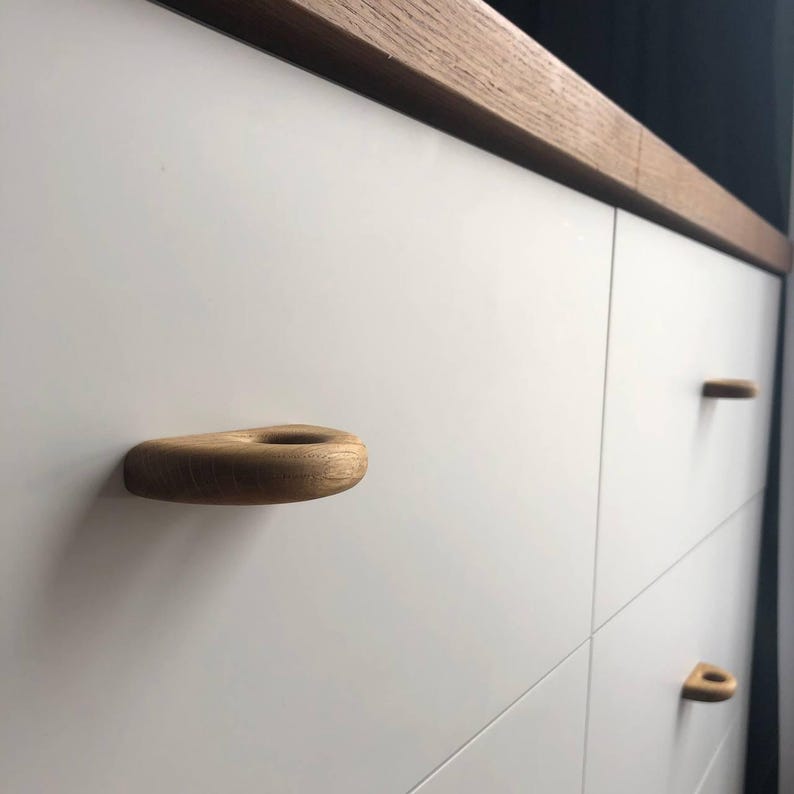 K&ouml;nnte beinhalten: Ein wei&szlig;er Schrank mit glatten, matten Oberfl&auml;chen und einer oberen Kante aus nat&uuml;rlichem, hellbraunem Holz. Die Schubladen des Schranks sind mit abgerundeten, tropfenf&ouml;rmigen hellbraunen Holzgriffen versehen, die eine mittige kreisf&ouml;rmige Aussparung aufweisen.