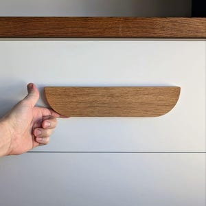 May include: A wooden drawer pull with a rounded, half-circle shape. The pull is made of light brown wood and is mounted on a white surface.