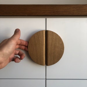 Pode incluir: Um close-up de uma porta de armário branca com uma maçaneta de armário de madeira em forma de semicírculo. A maçaneta é feita de duas peças de madeira que se juntam no meio.