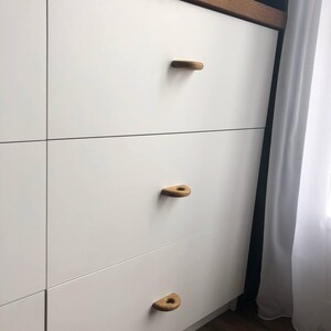 K&ouml;nnte beinhalten: Eine wei&szlig;e Kommode mit drei &uuml;bereinanderliegenden Schubladen, die jeweils mit einem halbkreisf&ouml;rmigen, hellbraunen Holzgriff versehen sind. Das M&ouml;belst&uuml;ck hat eine glatte, matte wei&szlig;e Oberfl&auml;che und eine dunkle Holzkante an der Oberseite.