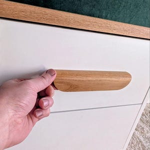 May include: A white cabinet with a light wood trim and a matching wooden handle. The handle is being held by a person. The cabinet is set against a dark green backdrop.
