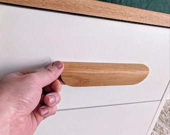 May include: A white cabinet with a light wood trim and a matching wooden handle. The handle is being held by a person. The cabinet is set against a dark green backdrop.