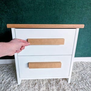 May include: A white nightstand with two drawers and a light wood top and drawer pulls. The nightstand is set against a dark green background and sits on a white rug.