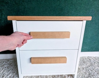 May include: A white nightstand with two drawers and a light wood top and drawer pulls. The nightstand is set against a dark green background and sits on a white rug.