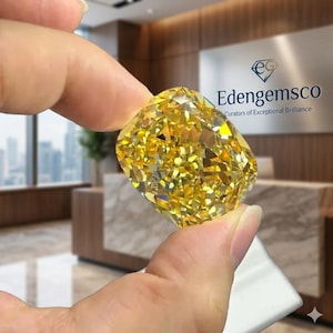 Op de afbeelding: Een grote, stralende gele diamant met een kussensnede wordt tussen vingers gehouden. De edelsteen schittert met talloze facetten en reflecteert licht. Het logo "Edengemsco, Curators of Exceptional Brilliance" is zichtbaar op de achtergrond.